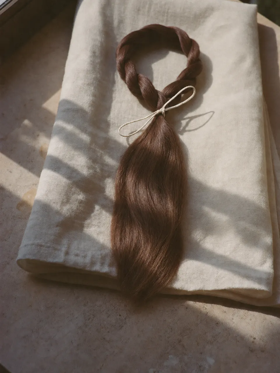 Mecha de cabelo castanho sedoso, amarrada com um laço de algodão, sobre tecido de linho à luz da janela